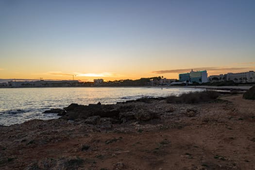 Scenic sunset over the rocky coastline in Mallorca, Spain. The golden light reflects on the calm sea, with buildings and palm trees silhouetted in the background.