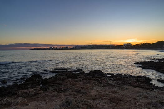 Scenic sunset over the rocky coastline in Mallorca, Spain. The golden light reflects on the calm sea, with buildings and palm trees silhouetted in the background.