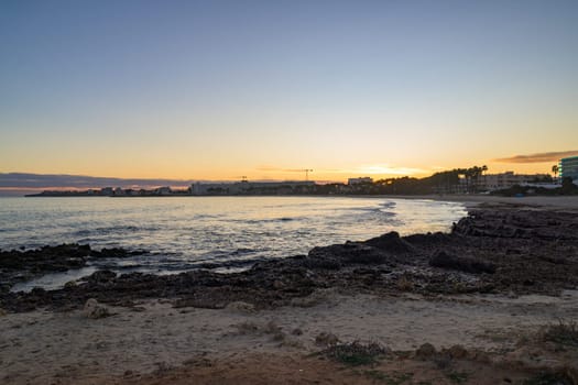 Scenic sunset over the rocky coastline in Mallorca, Spain. The golden light reflects on the calm sea, with buildings and palm trees silhouetted in the background.
