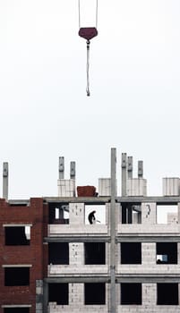 Amidst the backdrop of an overcast sky, construction workers diligently engage in the building of a large structure. A towering crane hovers above, carefully lifting construction materials to the upper levels of the framework. Below, men are seen maneuvering through the partially completed walls, ensuring precision as they install blocks.