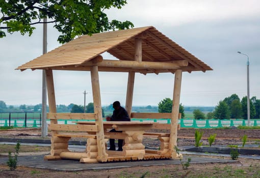 A person sits at a wooden picnic table in a gazebo, enjoying tranquility in a rural area with lush greenery.