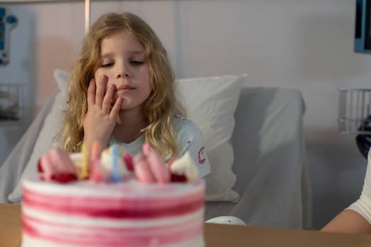 A young girl looks at a beautifully decorated birthday cake with longing in a hospital room setting