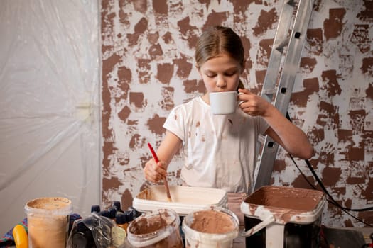 A young girl is creatively painting while holding a cup of her favorite drink in hand, showcasing her artistic talent