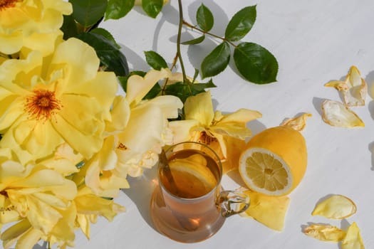 glass of tea with lemon and bouquet of yellow roses next to it on table with scattered rose petals creating natural still life on white table in the rays of the evening sun,