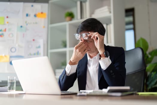 An elderly businessman rubs his eyes in frustration while working on a laptop, reflecting the pressures of modern business.