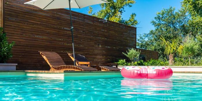 Modern backyard pool with turquoise water, pink inflatable ring, wooden deck, umbrella, and lounge chairs