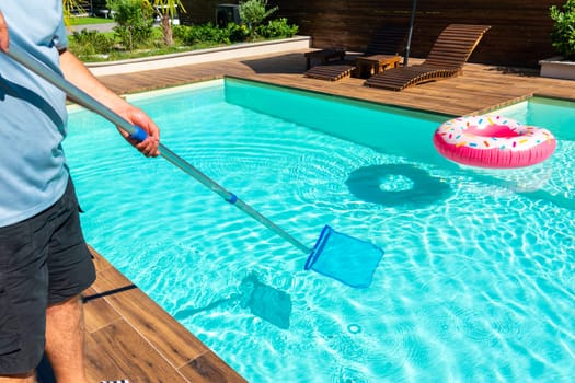 Man cleaning turquoise swimming pool with skimmer net, wooden deck, and colorful inflatable rings