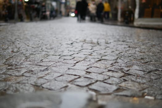 Wet cobblestones glisten as people walk in a busy urban area.