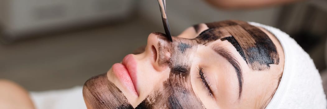 cosmetologist applying black mask on pretty woman face wearing black gloves, gorgeous woman in spa having facial procedures.