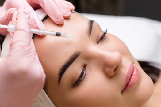 Close up of hands of young cosmetologist injecting botox in female face. She is standing and smiling. The woman is closed her eyes with relaxation.
