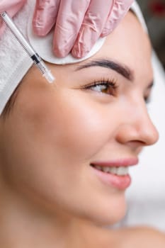 Close up of hands of young cosmetologist injecting botox in female face. She is standing and smiling. The woman is closed her eyes with relaxation.