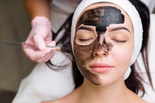 cosmetologist applying black mask on pretty woman face wearing black gloves, gorgeous woman in spa having facial procedures.