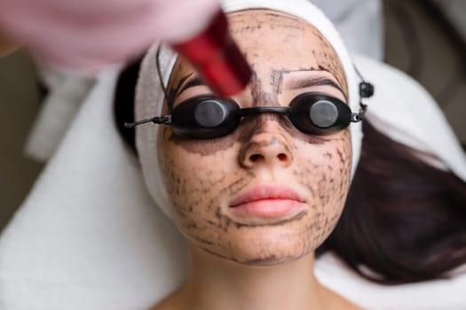Beautician doing hardware carbon peeling to a young beautiful client in a headdress in goggles lying on a sofa in a beauty salon. Facial cleansing concept. Cosmetology services.