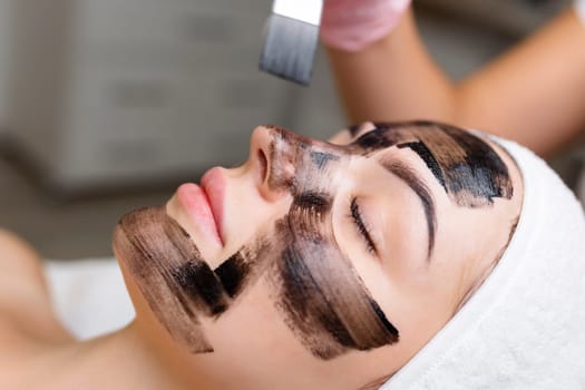 cosmetologist applying black mask on pretty woman face wearing black gloves, gorgeous woman in spa having facial procedures.