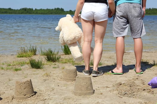 A happy couple enjoys a sunny beach day, building sandcastles while holding a teddy bear affectionately