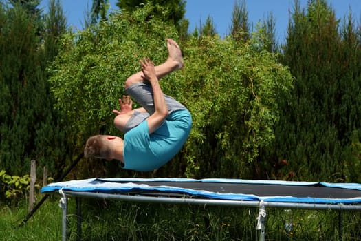 A young boy executes a truly impressive backflip while jumping on a trampoline in a bright sunny outdoor setting