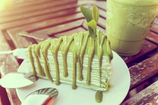 Vintage style soft tea leaf embroidered on matcha cake in white plate, placed near a glass of ice green tea on a old wooden table in the evening sunlight