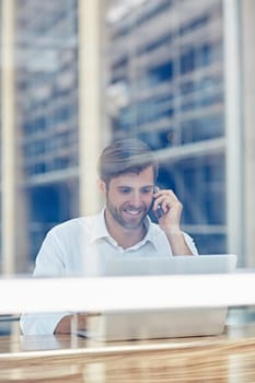 View through a window of a smiling businessman working on his laptop and talking on a mobile phone