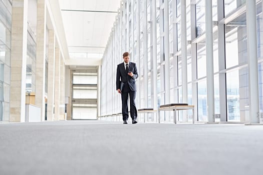 Low angle shot of a corporate executive walking along a modern corridor while using his mobile phone