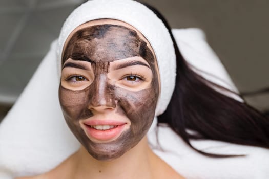 cosmetologist applying black mask on pretty woman face wearing black gloves, gorgeous woman in spa having facial procedures.