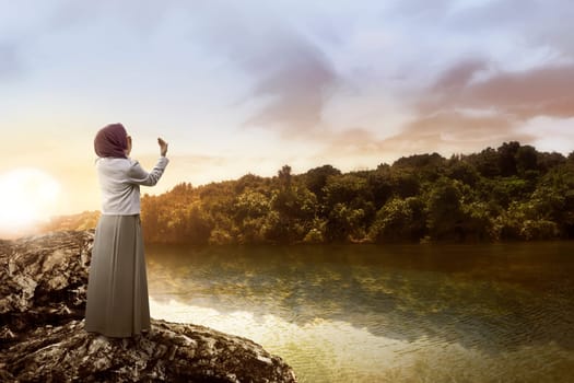 Young muslim woman praying standing on the rock at the lake
