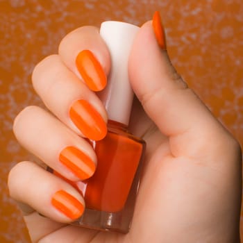 Female hand with orange nails holds orange nail polish on orange background.