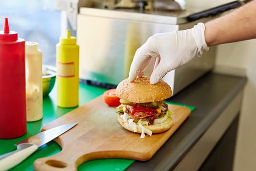Cropped shot of a white latex gloved hand carefully putting the top bun down on a big delicious burger that's been freshly prepared in a kitchen.