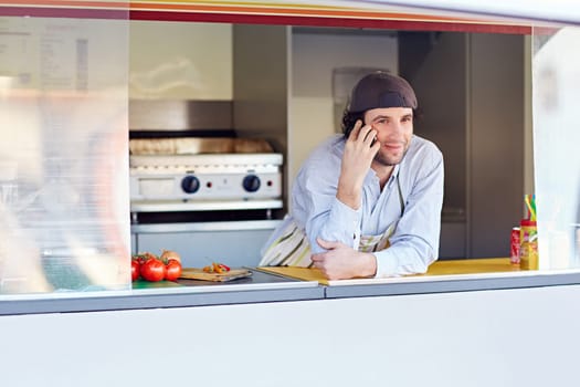 Male entrepeneur leaning on the counter and talking on the phone in his food stall where he makes and sells takeaway food