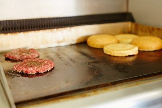 Two burger patties frying next to each other with open buns being toasted nearby.