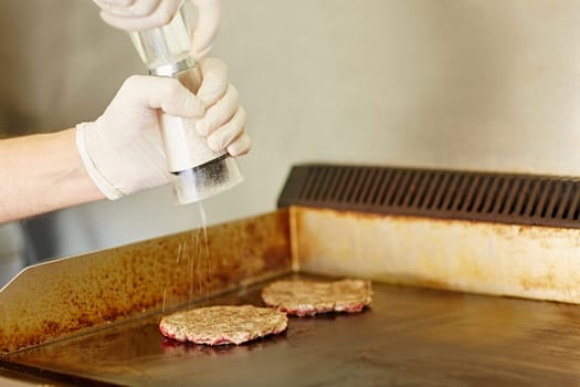 Cropped shot of gloved cook's hands grinding salt onto two hamburger patties to season them while they are frying on the grill