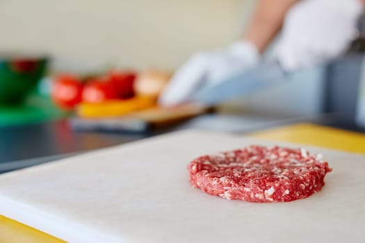 High quality raw hamburger patty sitting ready on a white board waiting for seasoning and cooking, while chef's hands prepare other ingredients in the background.