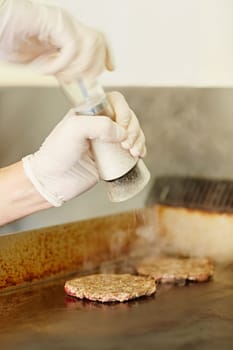 Cropped chef's hands wearing gloves busy grinding salt onto hamburger patties to season them while they are frying.
