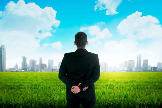 Asian business man standing on green meadow facing city. Business vision concept