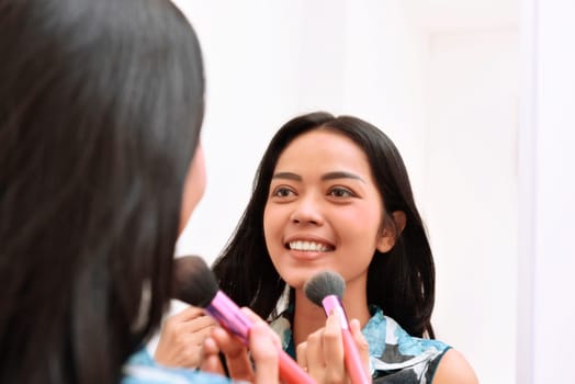 Beautiful asian girl applying makeup while looking on the mirror at home