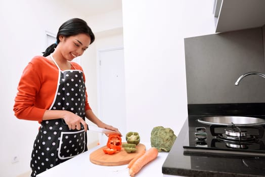 Smiling asian woman using knife to slice peppers and cooking in the kitchen