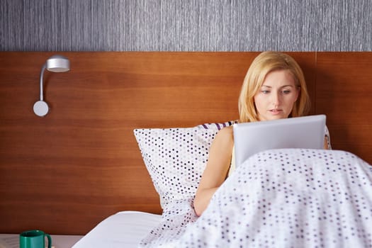 A young woman working on her tablet while lying in bed