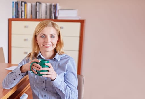 A young businesswoman smiling at the camera while sitting in her office drinking coffee