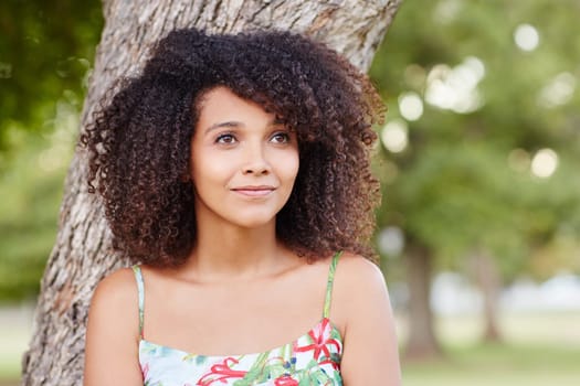 Young and beautiful mixed race woman smiling and leaning against a tree daydreaming in a green park