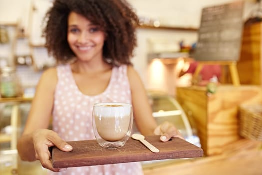 Focus on the foreground of a latte ay being carried on a wooden tray by a young mixed race waitress