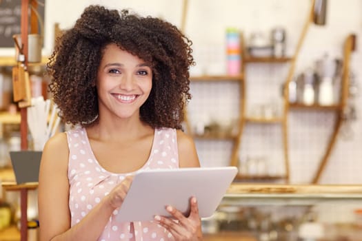 Portrait of a beautiful young woman with an afro hairstyle smiling at the camera while typing on her digital tablet in her coffee shop