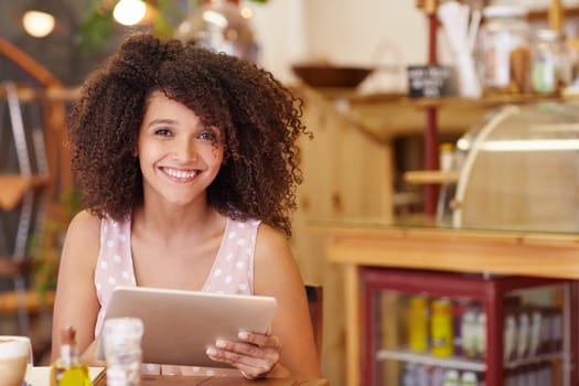 Beautiful young mixed race woman smiling at the camera confidently while using her digital tablet in a coffee shop