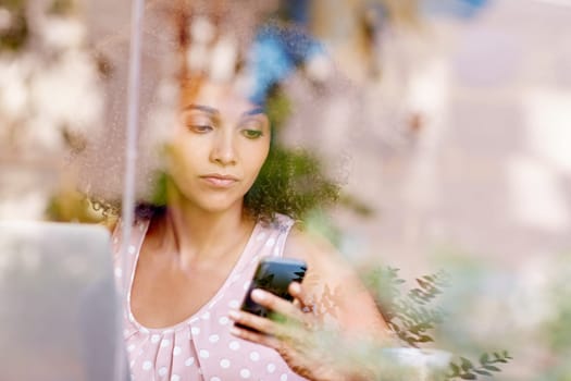Beautiful mixed race woman sitting in a coffee shop, seen through the window while typing on her mobile phone