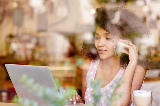 Beautiful mixed race woman talking on her phone while typing on her laptop, seen through the window of a coffee shop