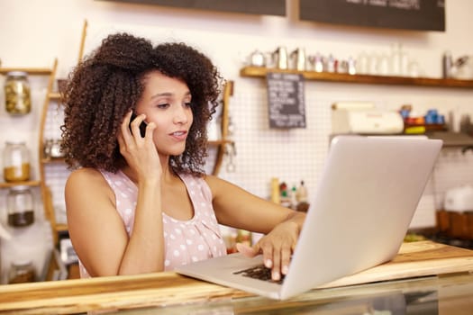 Small business owner in her coffee shop typing on her laptop while talking on the phone