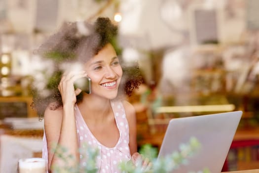Young mixed race woman seen through the window of a coffee shop smiling and talking on her mobile phone with her laptop open