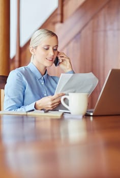 A young businesswoman at her desk, talking on phone while checking documents, inside wooden office