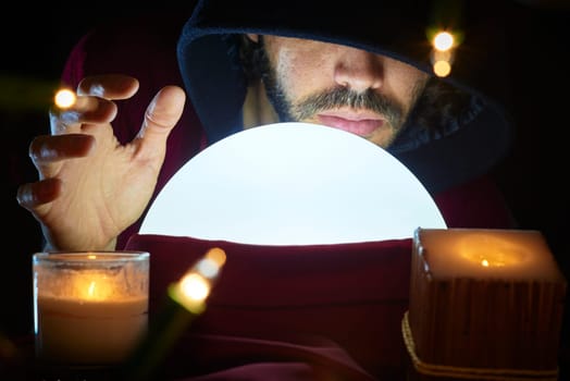 Very low key portrait of hooded man with eyes covered reading fortune on bright crystall ball, surrounded by candle light.