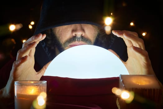 Very low key portrait of hooded man with eyes covered reading fortune on bright crystall ball, surrounded by candle light.
