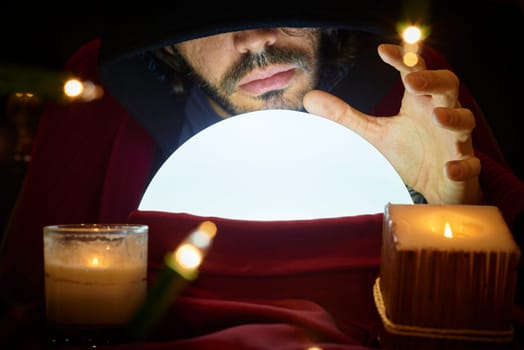 Very low key portrait of hooded man with eyes covered reading fortune on bright crystall ball, surrounded by candle light.