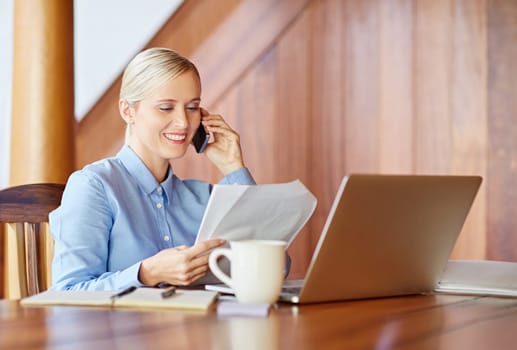 A young businesswoman at her desk, talking on phone while checking documents, inside wooden office